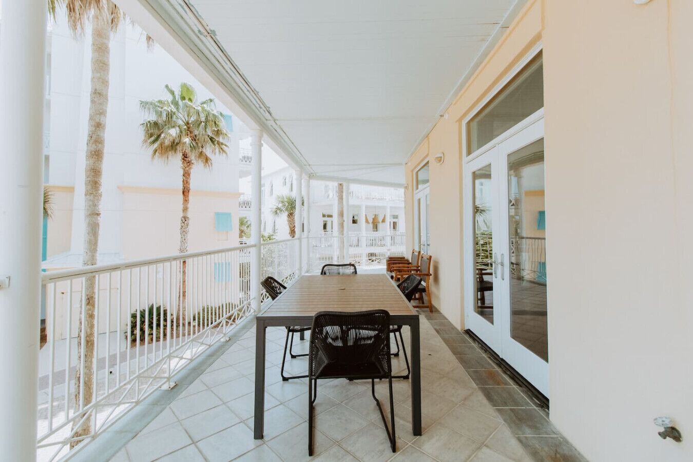 A covered balcony with a wooden table and chairs, palm trees, and a view of other buildings.