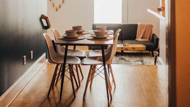 A modern dining area with a wooden table and chairs, a black wall with a white ceiling vent, and a view into a living space with a sofa and a window. - Historic Downtown Loft (Waxahachie)