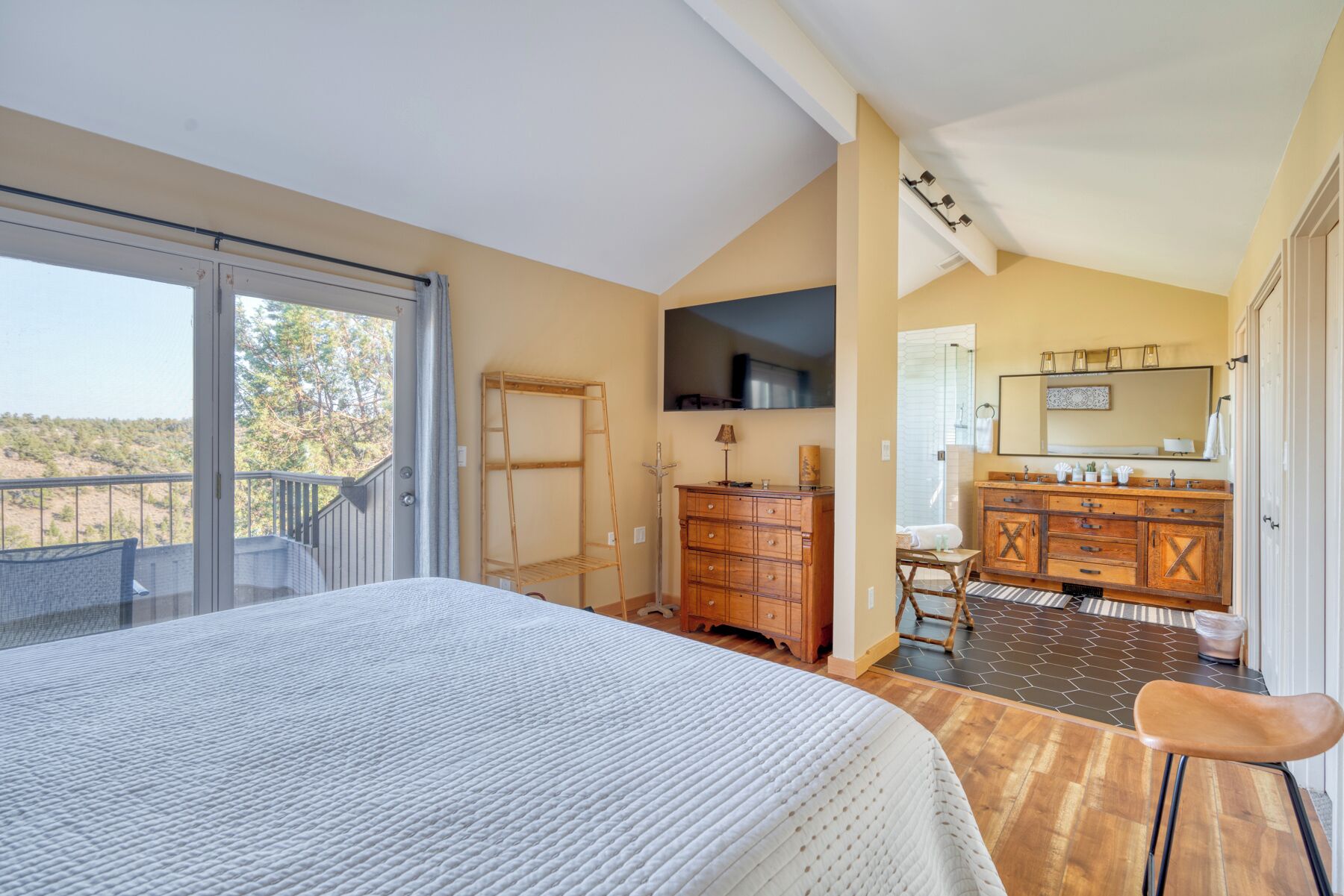A bedroom with a large bed, a wooden dresser, a vanity with a mirror, and a balcony with a view of the landscape.