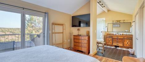 A bedroom with a large bed, a wooden dresser, a vanity with a mirror, and a balcony with a view of the landscape.