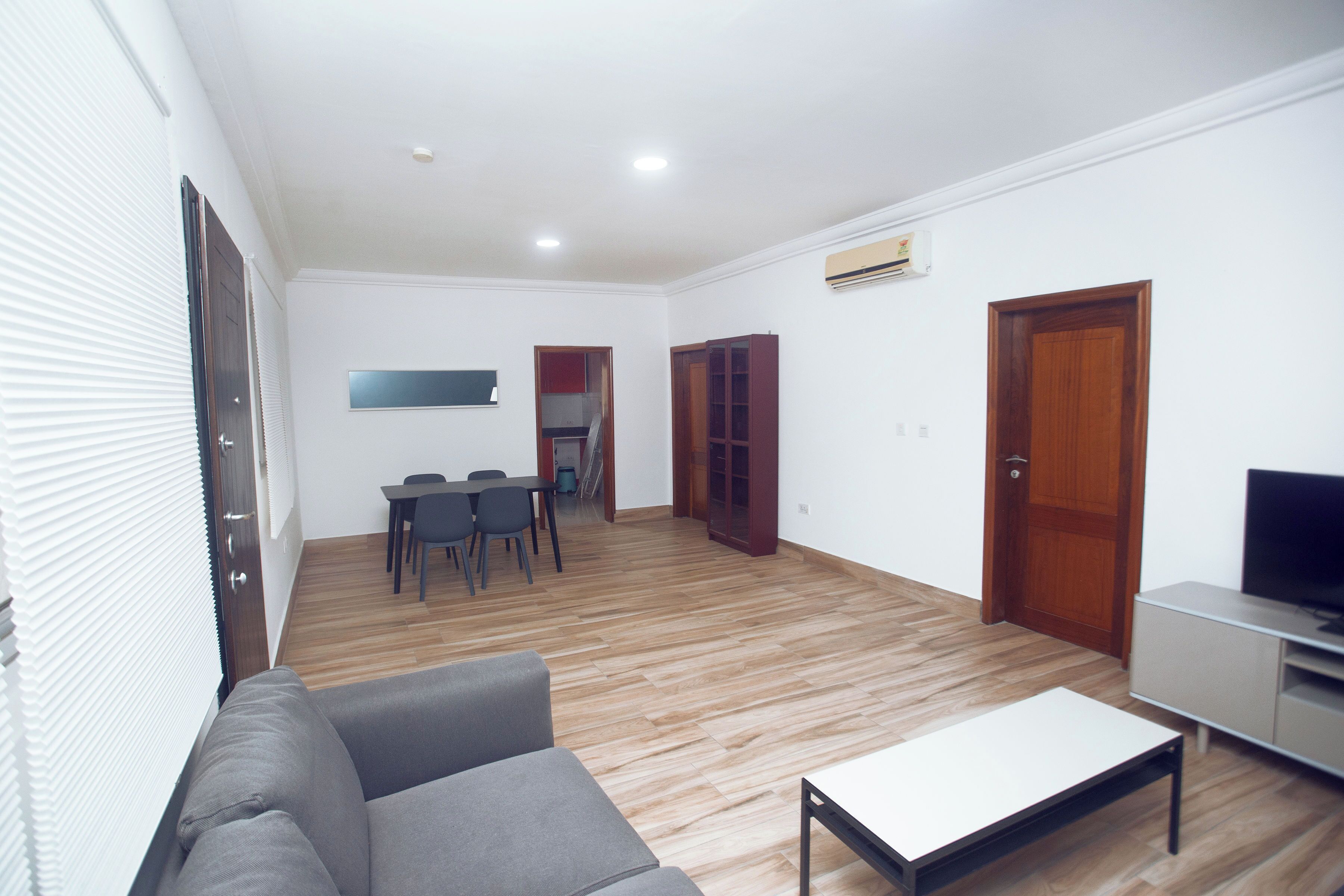 A modern living room with a grey sofa, a coffee table, and a TV stand. There is a dining area with a table and chairs, and a kitchen area with cabinets and a sink.