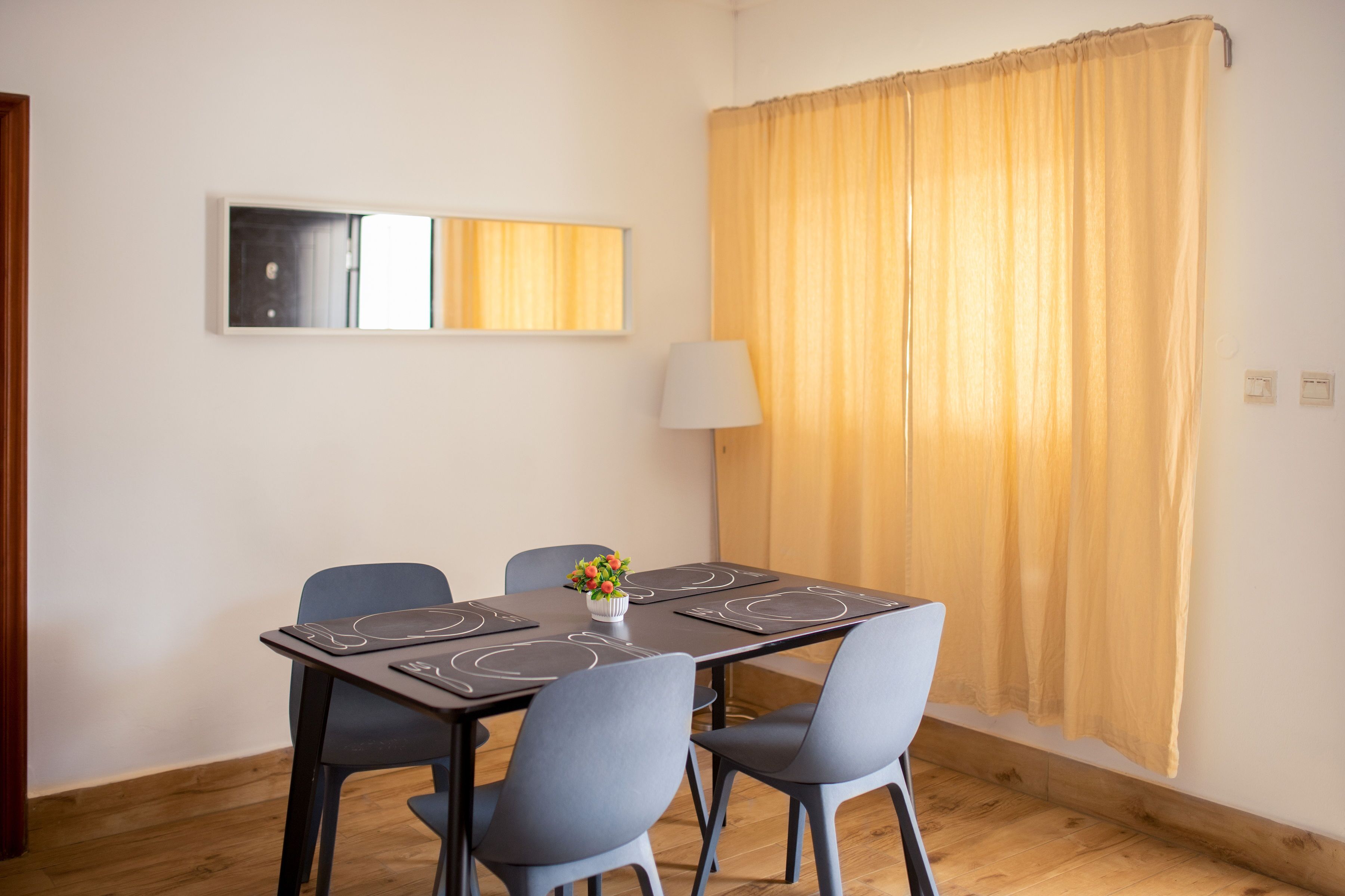 Un coin repas avec une table dressée pour quatre personnes, un miroir au mur et une lampe près de la fenêtre.