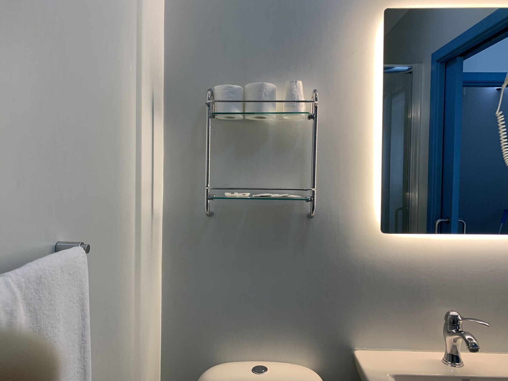 Un baño con repisas de vidrio que sostienen artículos de tocador, un lavamanos blanco y un espejo.