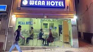 The exterior of WEAA HOTEL with a glass facade, outdoor seating area, and a visible entrance. - WEAA Hotel (Nazaré)