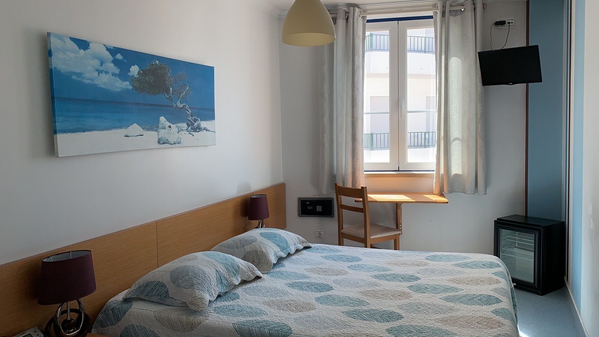 A bedroom with a bed, a desk, a chair, a TV, and a painting on the wall.