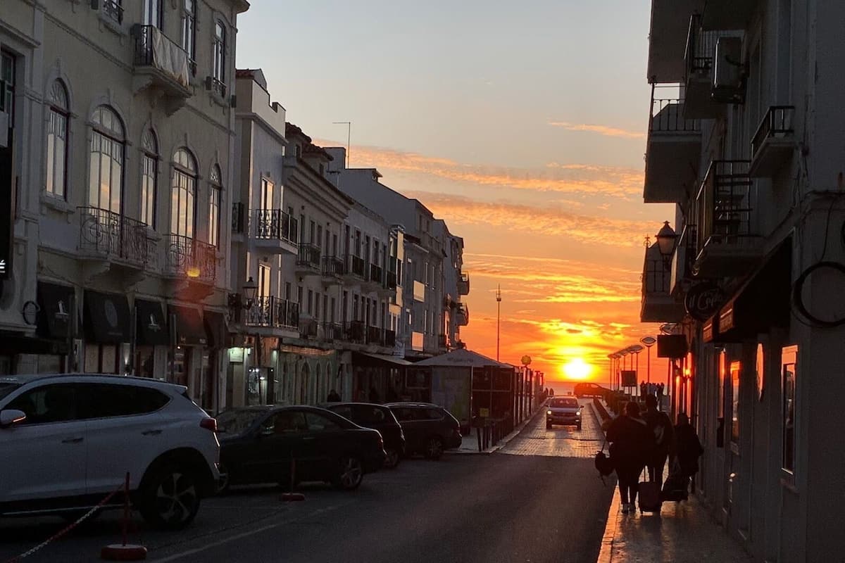 A street at sunset with buildings on either side, a car parked on the left, and a person walking on the sidewalk.