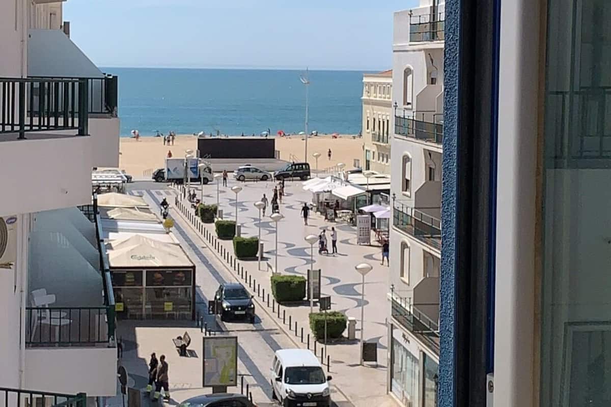 A view from a window overlooking a street with parked cars, a beach, and buildings.