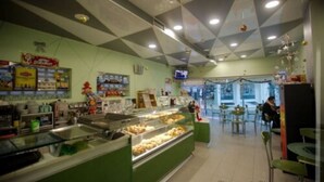 A bakery with a green counter displaying pastries, a seating area with tables and chairs, and a ceiling with geometric patterns. - WEAA Hotel (Nazaré)