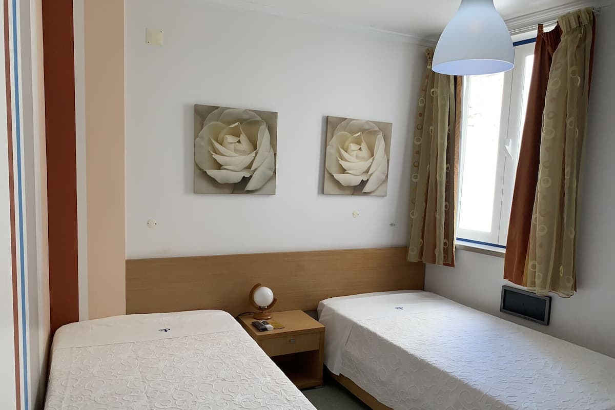 A hotel room with two single beds, a wooden headboard, a bedside table with a lamp, and two large floral paintings on the wall.