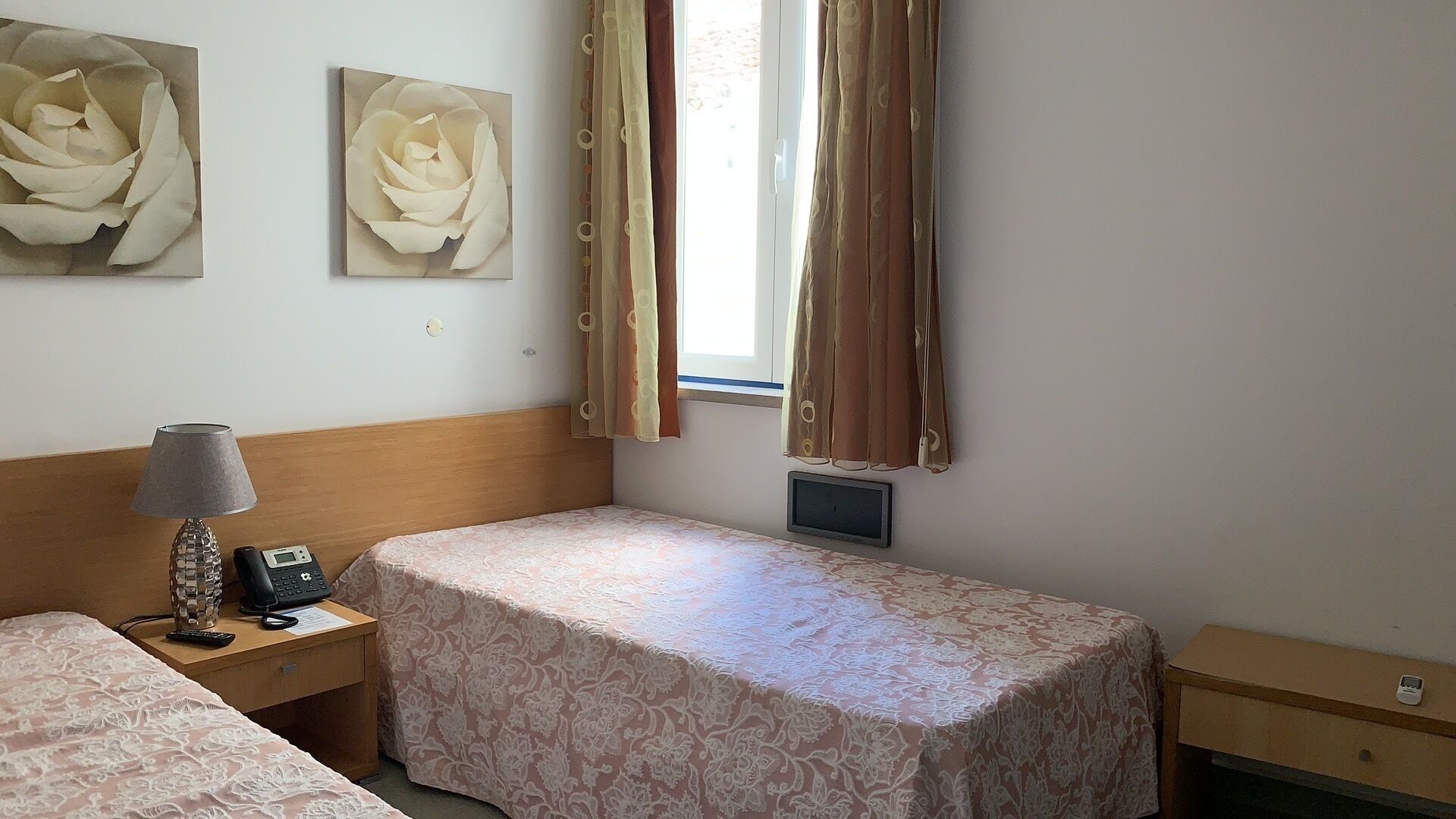 Une chambre d’hôtel avec un lit simple, une table de chevet avec une lampe et un téléphone, une fenêtre avec des rideaux et deux tableaux représentant des fleurs, accrochés au mur.