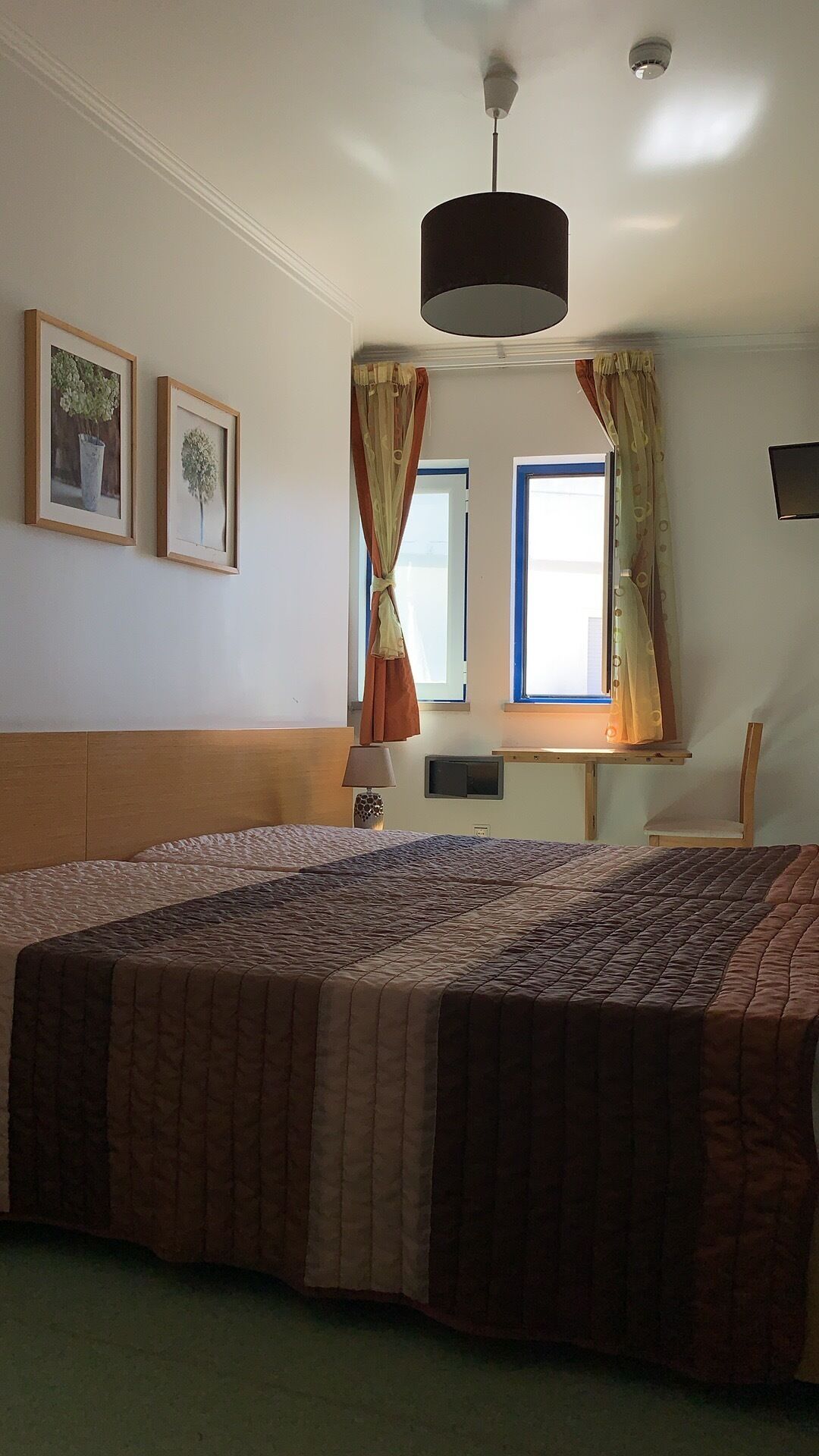 A bedroom with a large bed, a window with curtains, a ceiling light, and framed pictures on the wall.