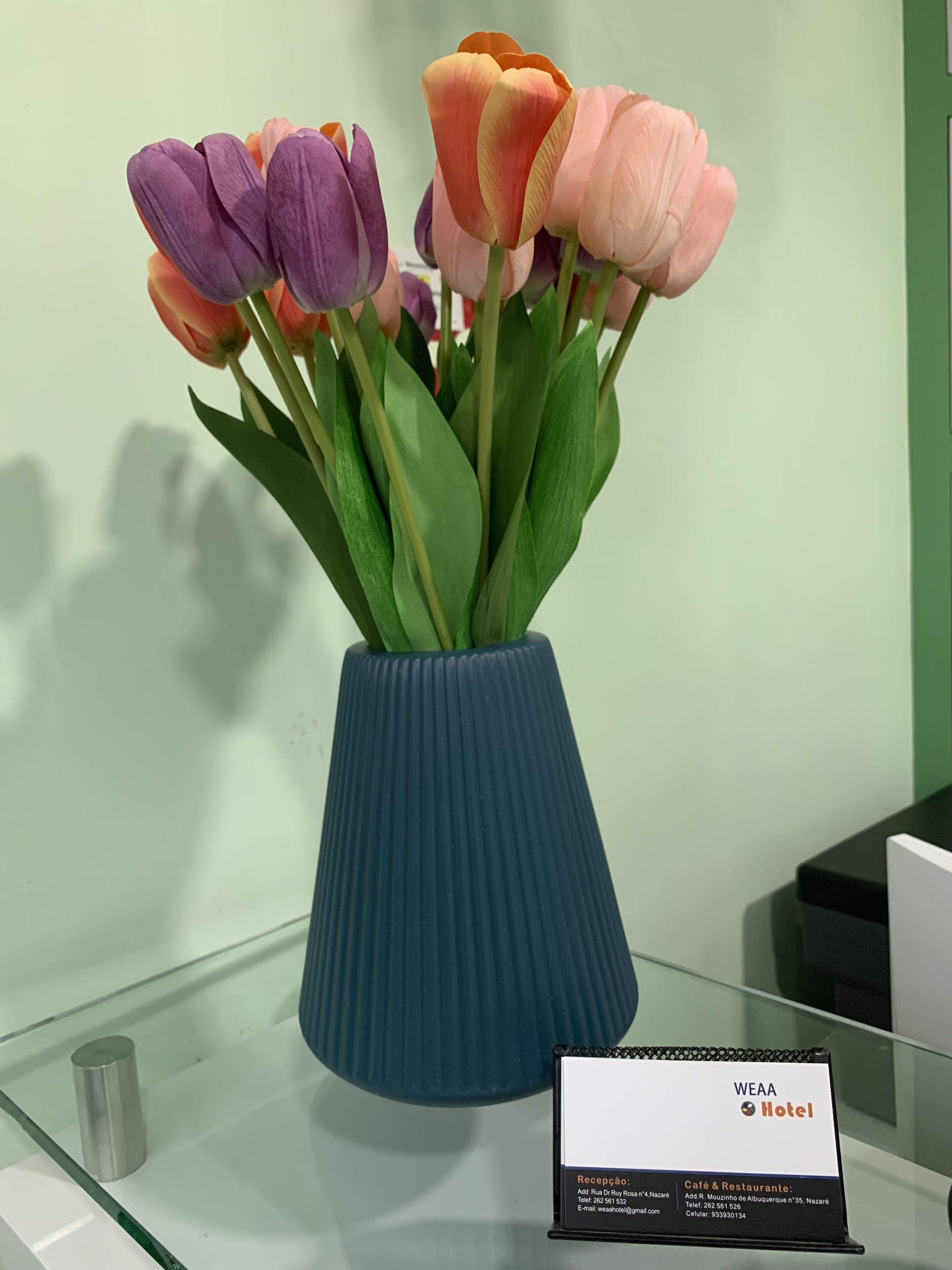 Un jarrón azul con tulipanes sobre una mesa de vidrio. Se ve un cartel con información del hotel.