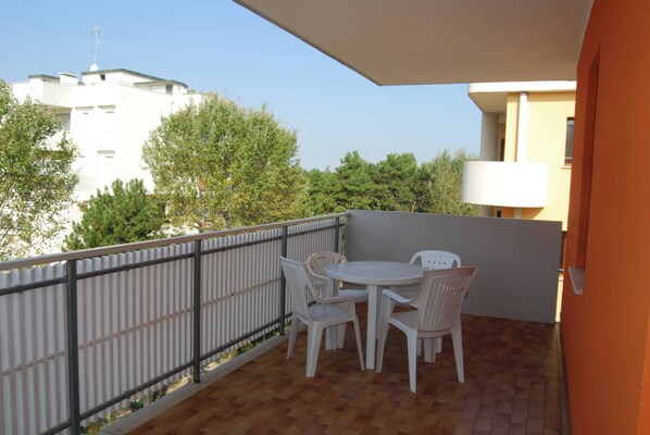 Un balcon avec une table et des chaises blanches, une rambarde en métal et une vue sur les arbres et les bâtiments.