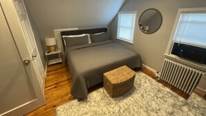 Un dormitorio con cama, mesita de noche, lámpara, ventana, radiador y una puerta.
