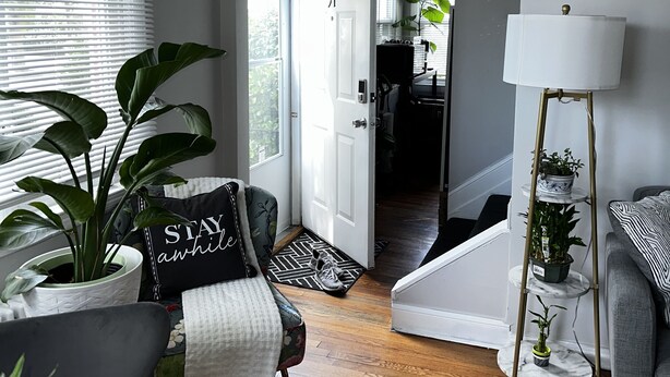 Una sala moderna con un sofá gris, un sillón blanco, una mesa de centro y una lámpara. Hay una planta grande en maceta y una mesita con lámpara. Una puerta da a otra habitación.