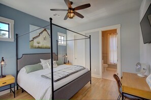 A four-poster bed with a ceiling fan, a bedside table with a lamp, and a painting of a house on the wall.