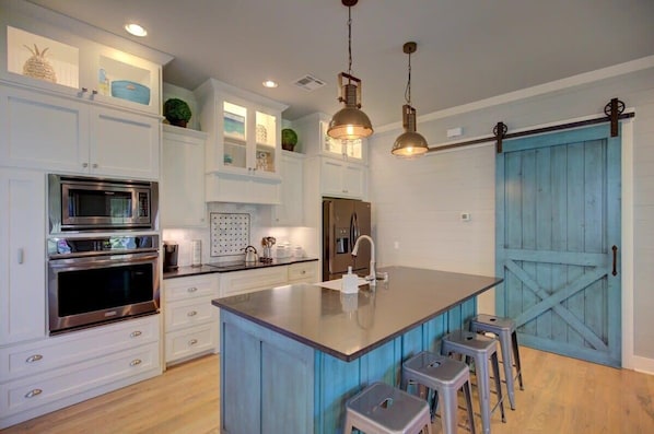 Una cocina moderna con armarios blancos, electrodomésticos de acero inoxidable, una isla central con taburetes y una puerta azul.
