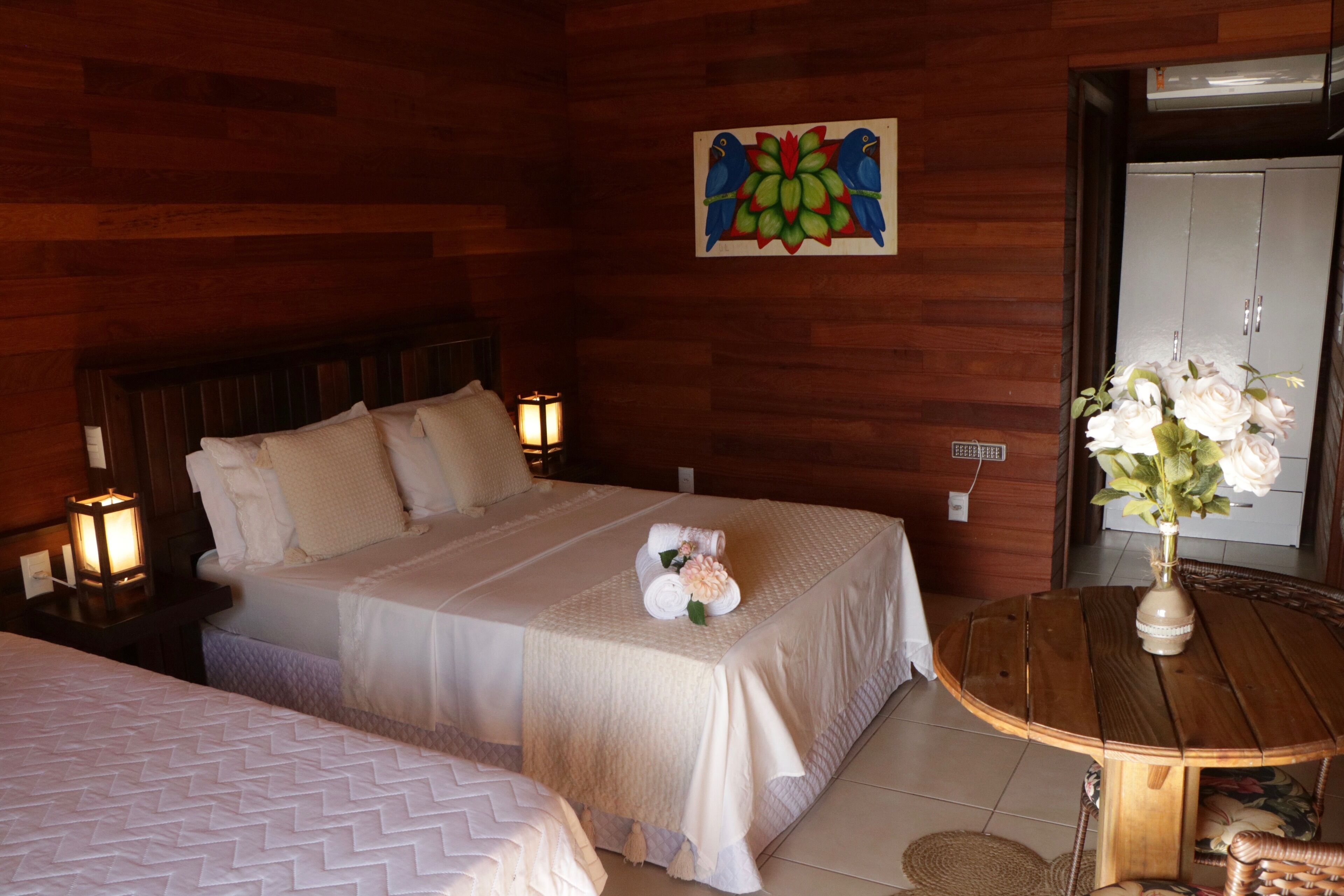 A hotel room with a wooden wall, a bed with white bedding, a nightstand with a lamp, and a round wooden table with a vase of flowers.