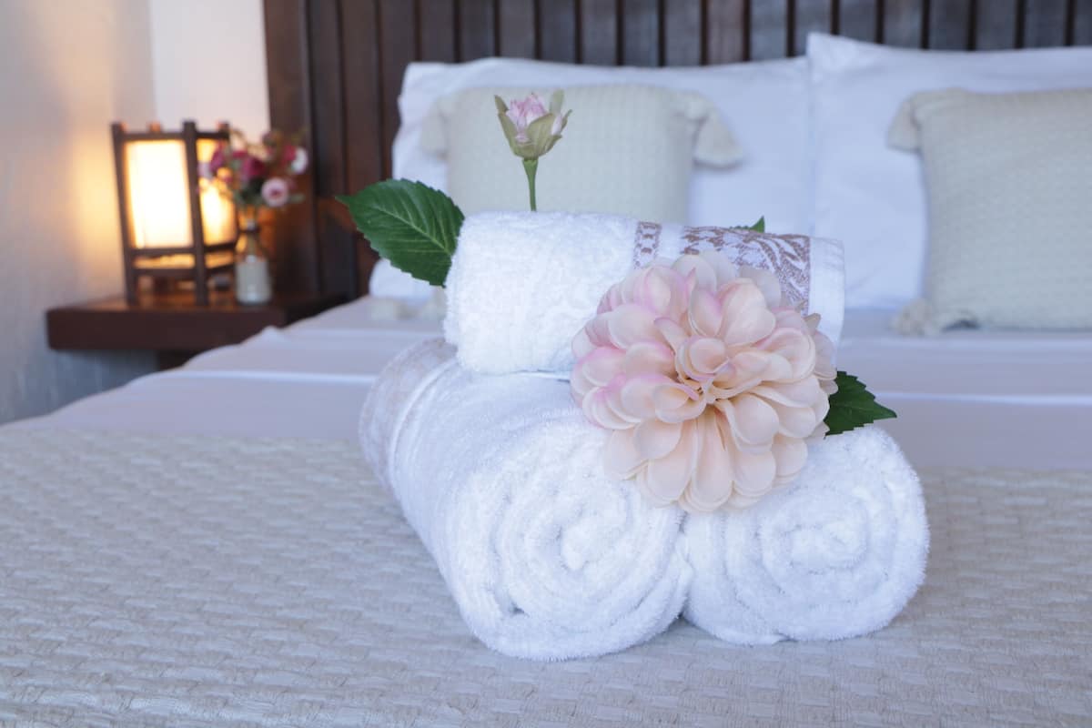 A neatly made bed with white linens, a decorative flower, and a rolled towel on a textured bedspread.