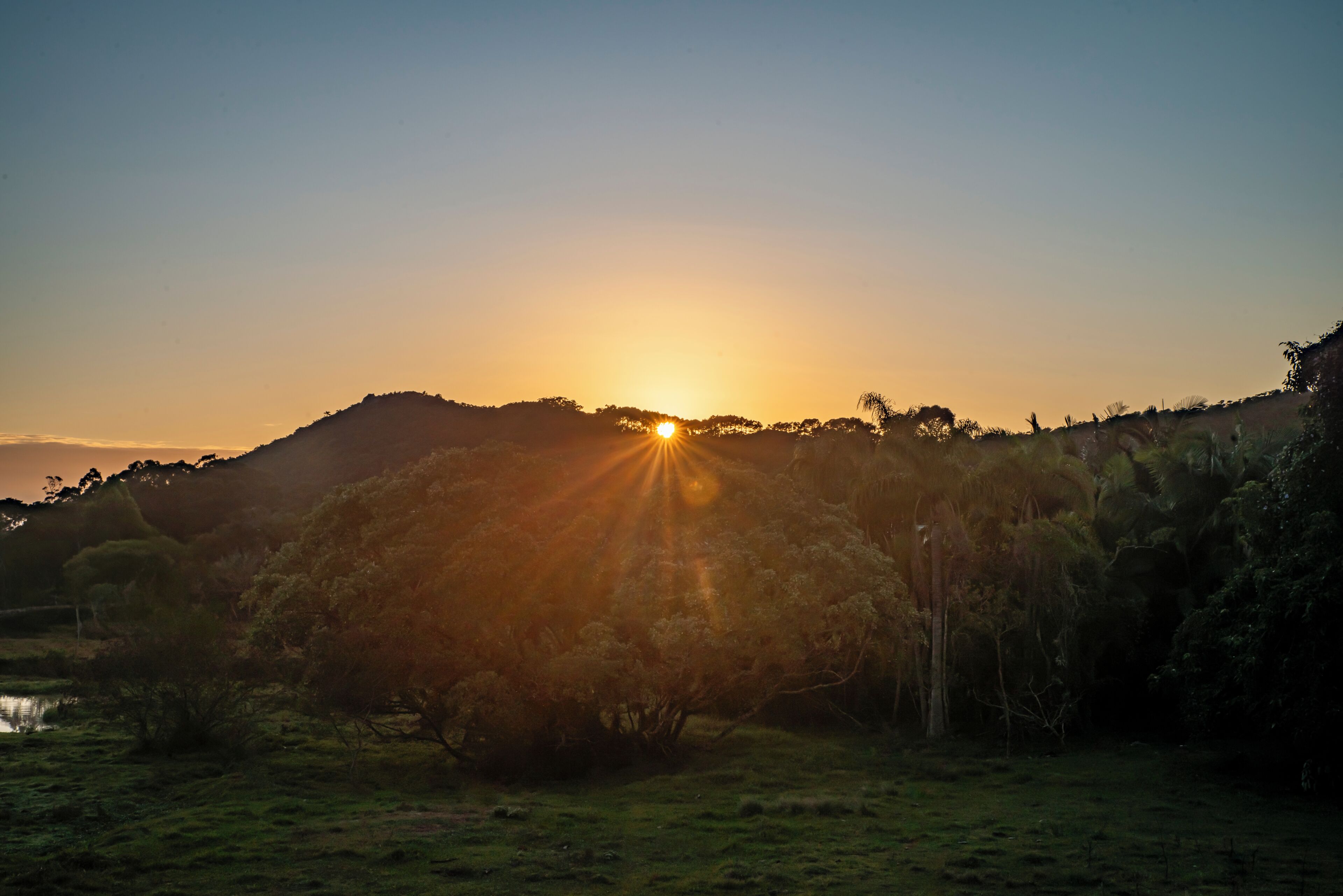 A sunset over a hilly landscape with trees and a small body of water.