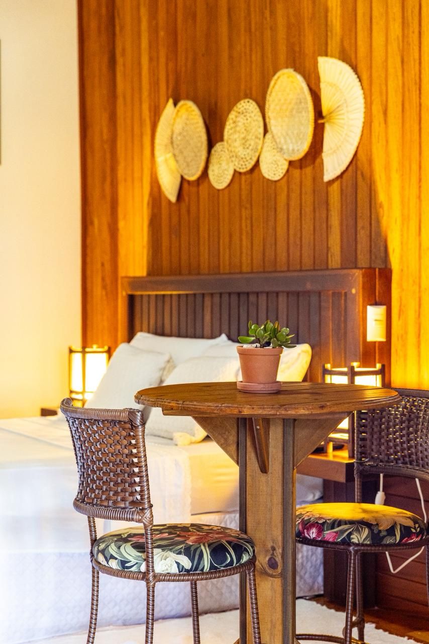 Una cama con estructura de madera y ropa de cama blanca, una mesa redonda de madera con una planta en maceta, y sillas de mimbre con cojines estampados.