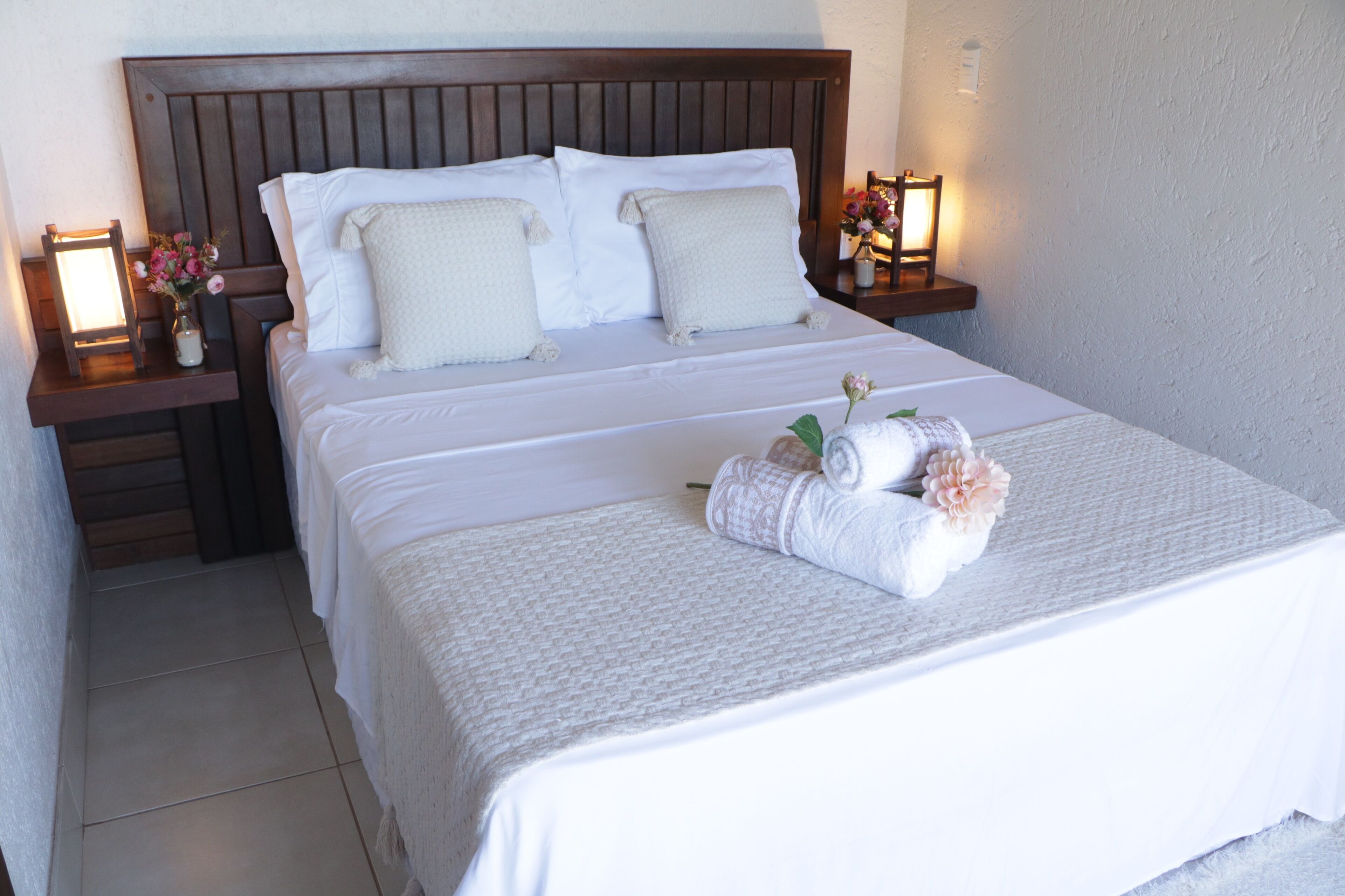Uma cama bem arrumada com lençóis e almofadas brancas, uma cabeceira de madeira e duas mesas de cabeceira com luminárias e flores.