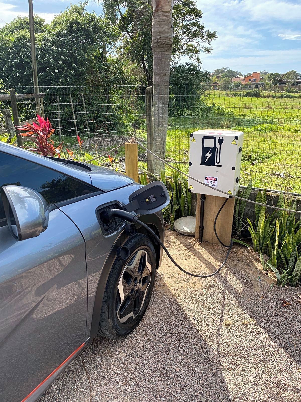 A car connected to an electric charging station in a rural setting with a fence and greenery in the background.