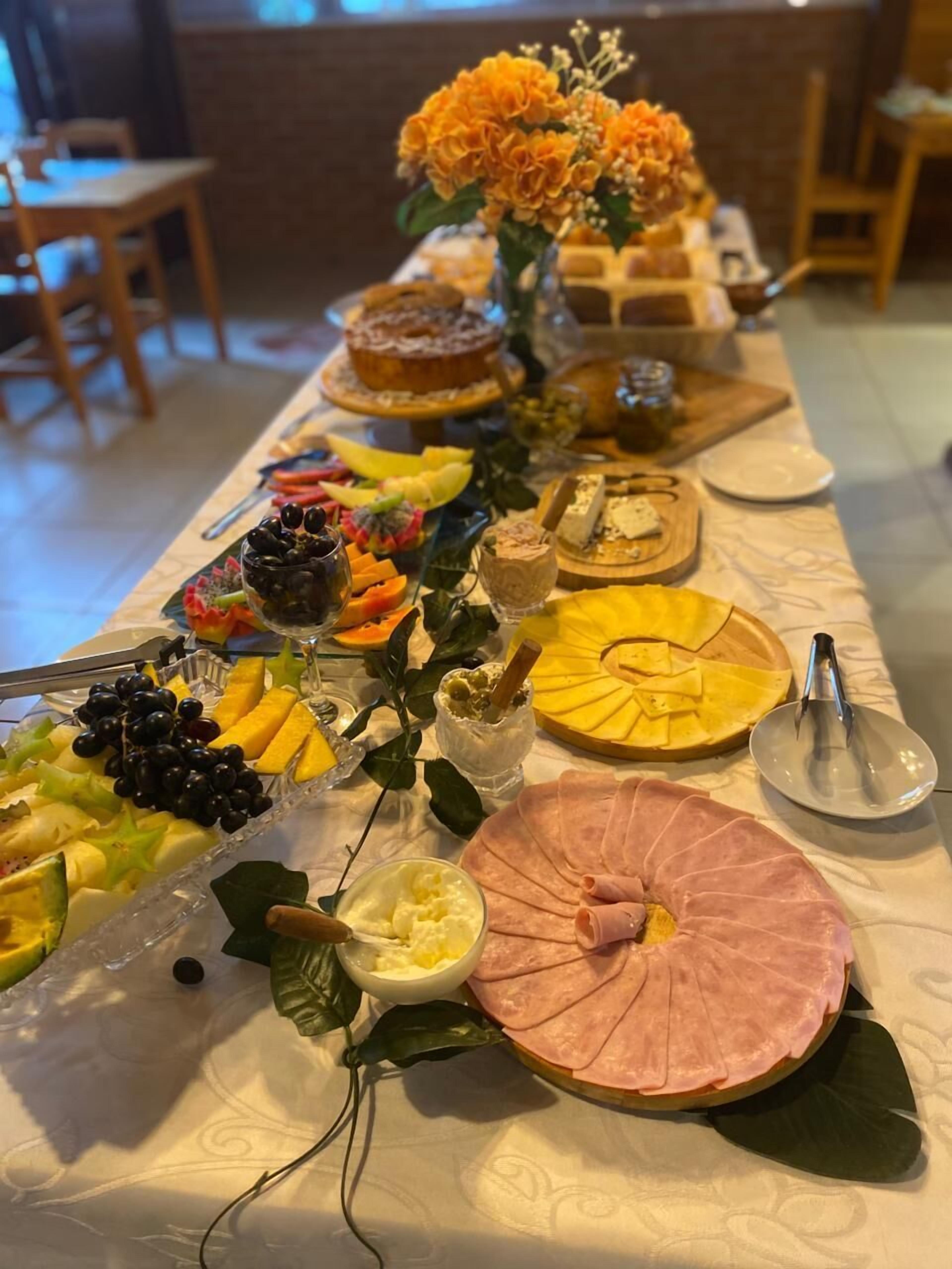Uma mesa de buffet com diversos alimentos, incluindo frutas, queijos e um presunto inteiro em rolo.