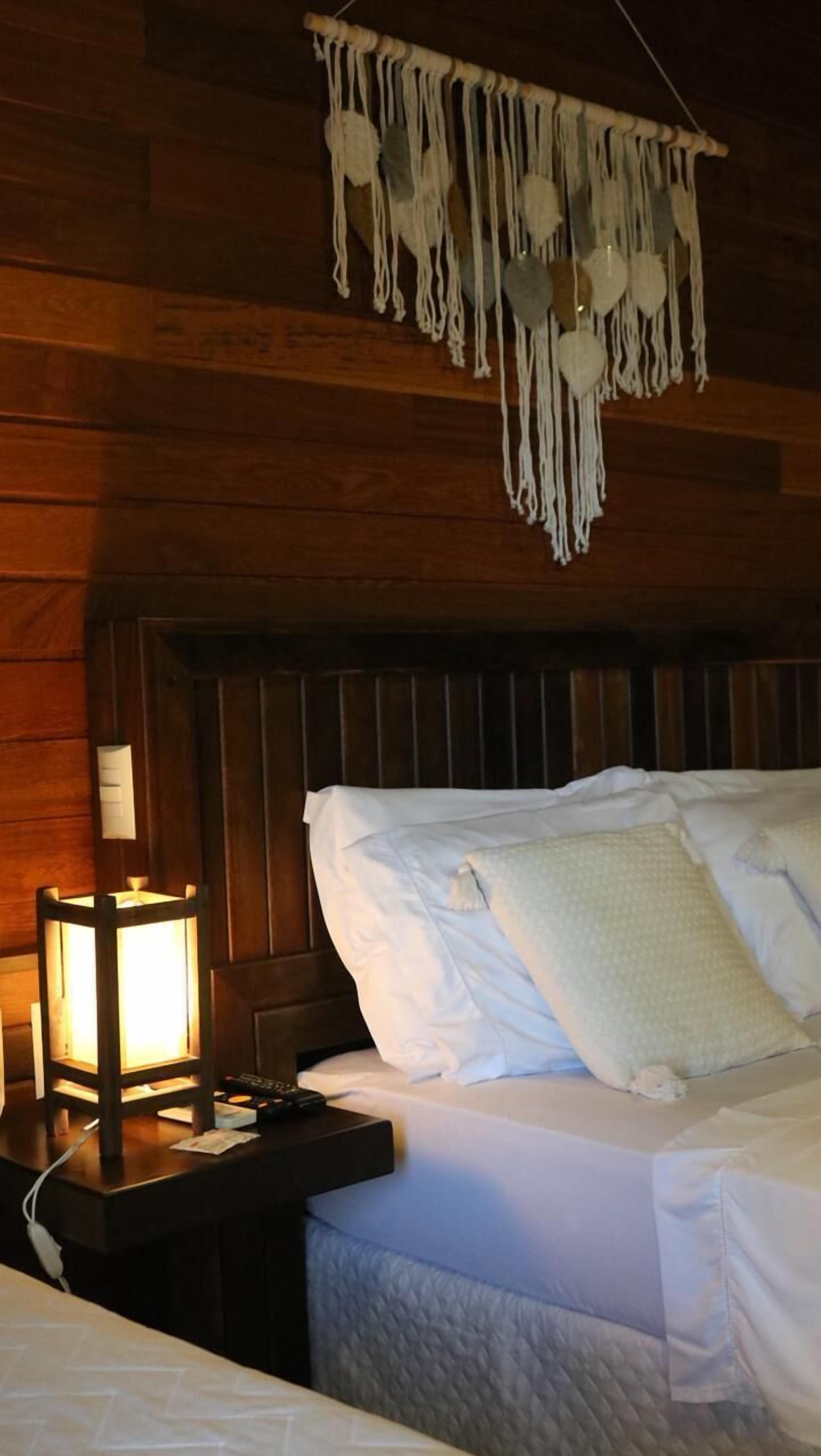 Habitación con paneles de madera, una cama, una mesita de noche, un tapiz de macramé en la pared y una lámpara de mesa encendida.