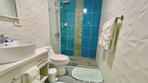 Un baño con lavamanos, inodoro y ducha blancos. Hay dos toallas en el suelo.