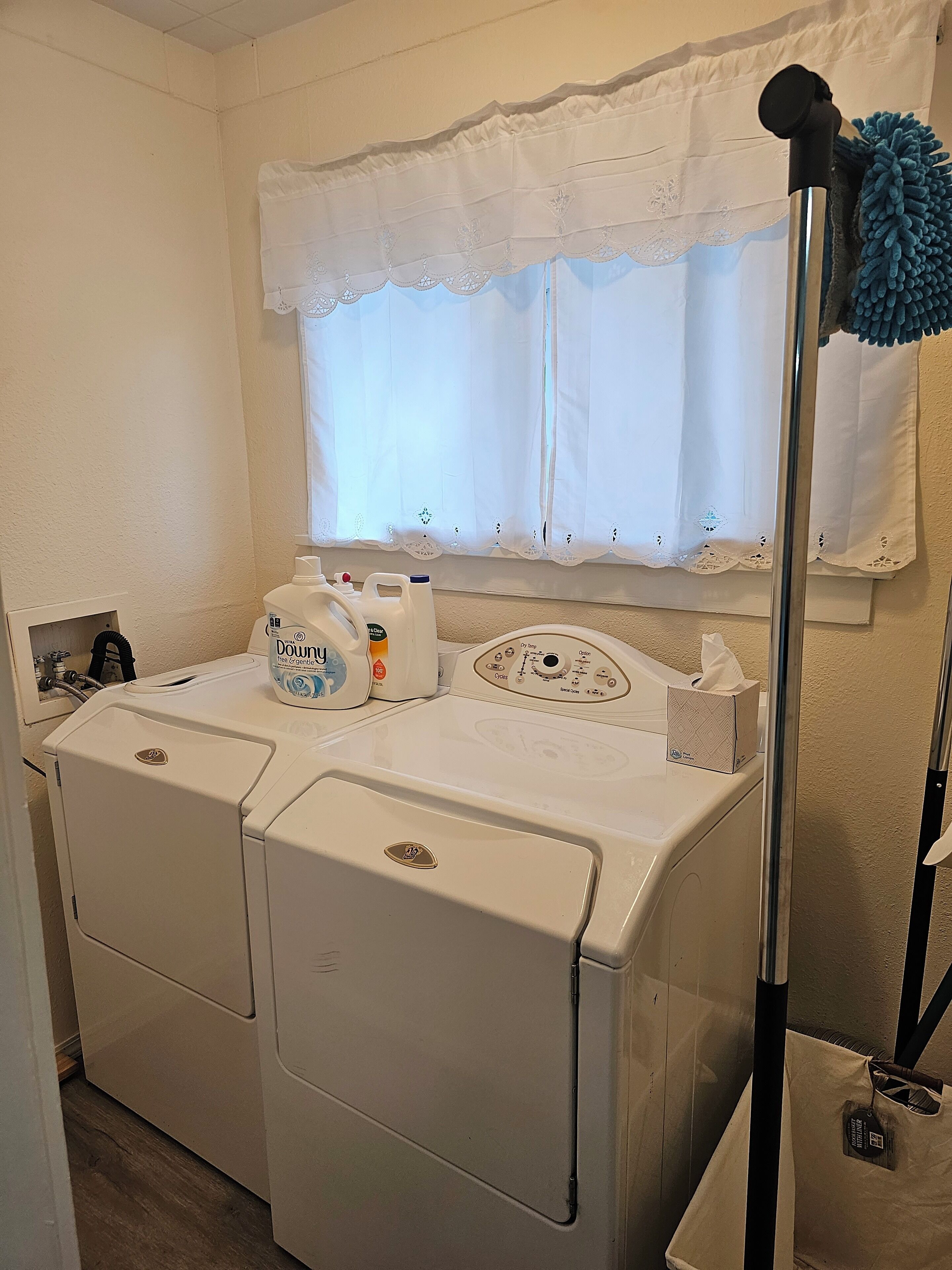Pesutila, jossa on etuaukollinen pesukone ja kuivausrumpu, ikkuna, jossa on pitsiverhot, ja sininen moppi nojaa seinää vasten.