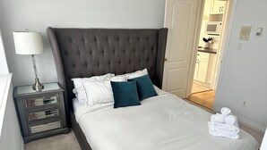 En pænt redt seng med hvide og teal farvede puder, en grå sengegavl og et natbord med lampe og skuffer.