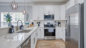 A modern kitchen with white cabinets, stainless steel appliances, a large island, and a view of the outdoors. - Beach Home! Private Pool! Arcade-Beach-FUN! Elevator (Murrells Inlet)