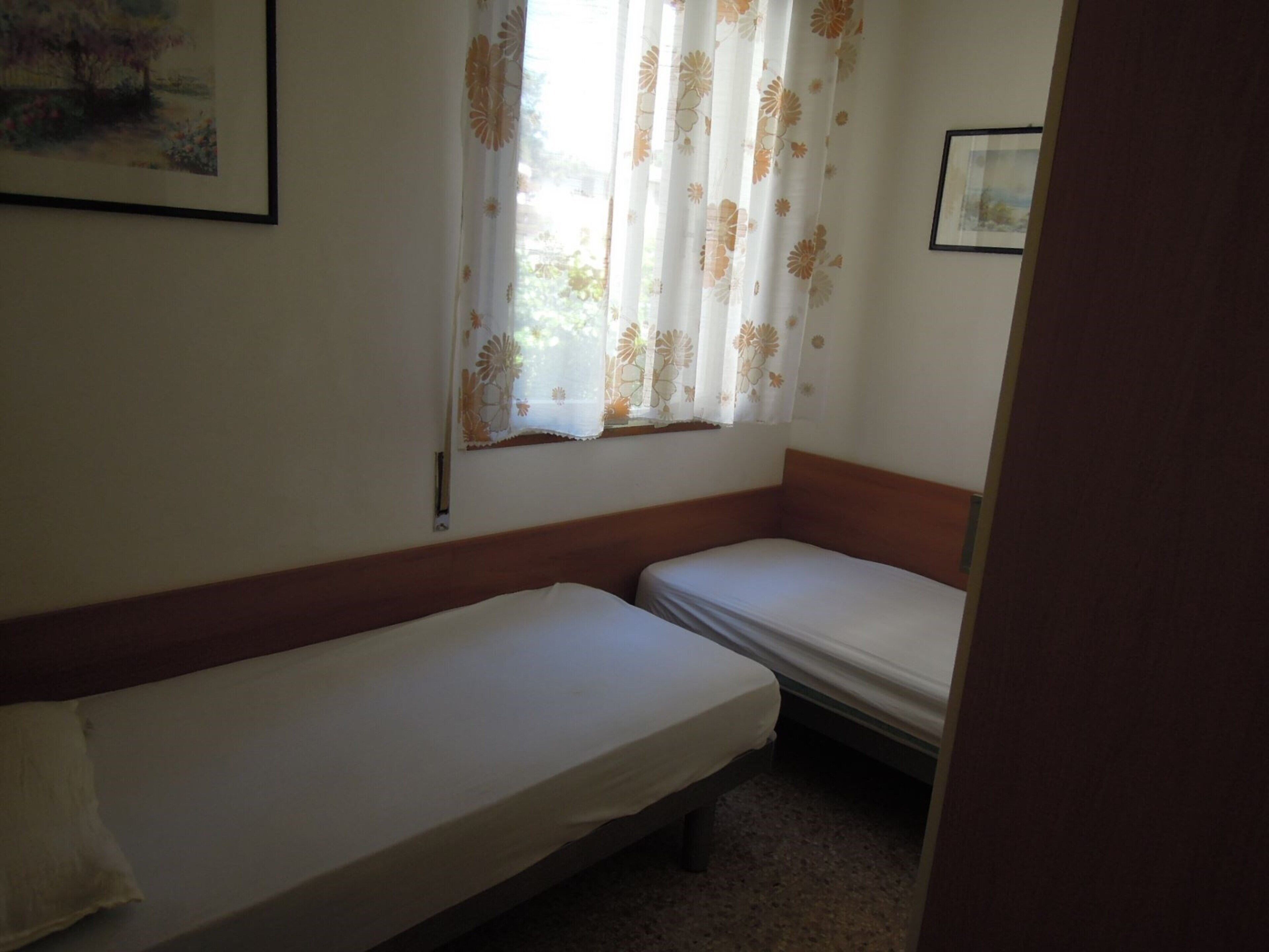 A small, single-room with two single beds, a window with floral curtains, and a framed picture on the wall.