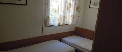 A small, single-room with two single beds, a window with floral curtains, and a framed picture on the wall.