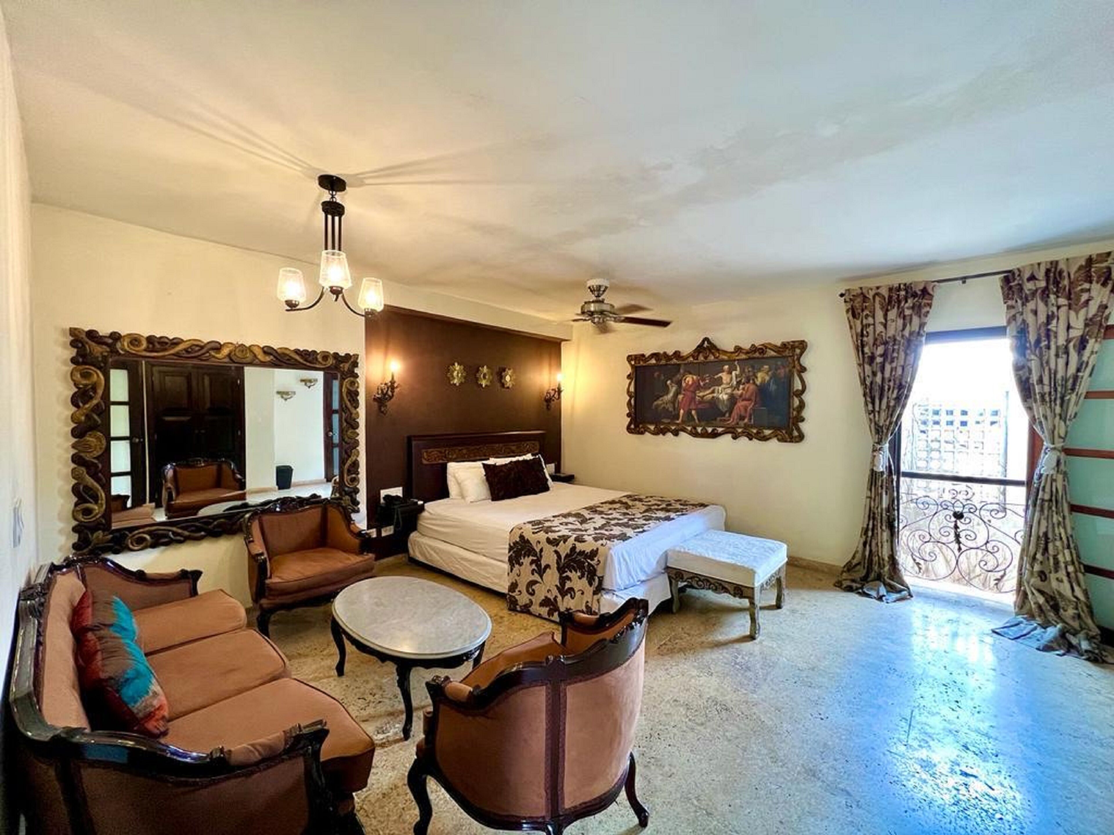 A hotel room with a large bed, a sitting area with a sofa and armchairs, a small round table, a ceiling fan, a large mirror, and a painting on the wall.