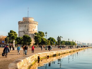 Una torre histórica de base circular y asta, rodeada por un paseo marítimo con árboles y una masa de agua que refleja la estructura.