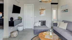 A modern living room with a grey sofa, a round wooden coffee table with glasses and a bottle, a mounted TV, and a large painting on the wall. - Sanders Old Port - Cozy 1-bdr. Apt. w/ Balcony (Thessaloniki)