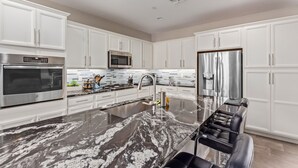 A modern kitchen with white cabinets, stainless steel appliances, a marble island, and a built-in hair station. - Gilbert Haven: A Family Delight in Enchanting Community (Gilbert)