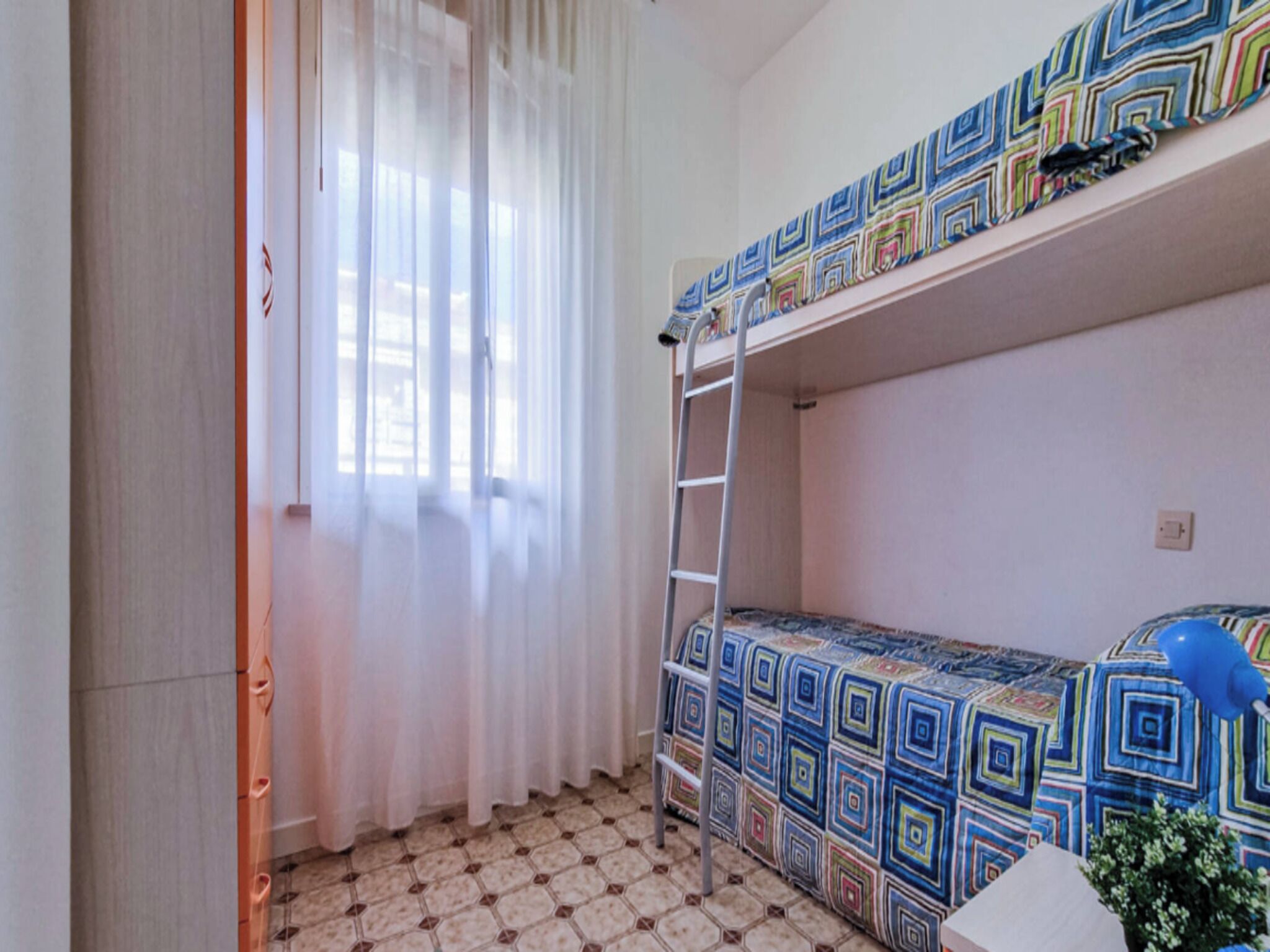 Ein Schlafsaalzimmer mit Etagenbetten, einer Stehleiter, einem Fenster mit Vorhängen und einem gefliesten Boden.