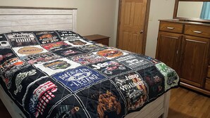 Un dormitorio con una cama cubierta con colcha de motivos Jeep, una puerta de madera, un aparador y un equipo de aire acondicionado blanco en la pared.