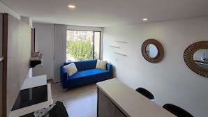 A modern living room with a blue sofa, a white coffee table, and a flat-screen TV. There is a large window with a view of buildings and trees. - Rolo Apartamentos Amoblados Jerez (Bogotá)