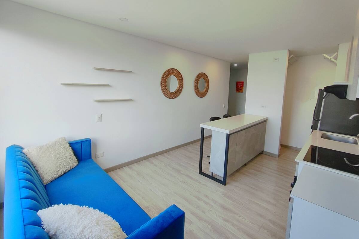 A modern living room with a blue sofa, a small white table, and a kitchen area with a white countertop and cabinets.