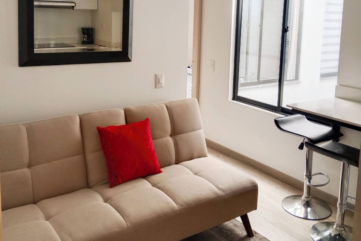 A modern living room with a beige sofa, a red pillow, a black-framed mirror, a small kitchen area, and a large window.