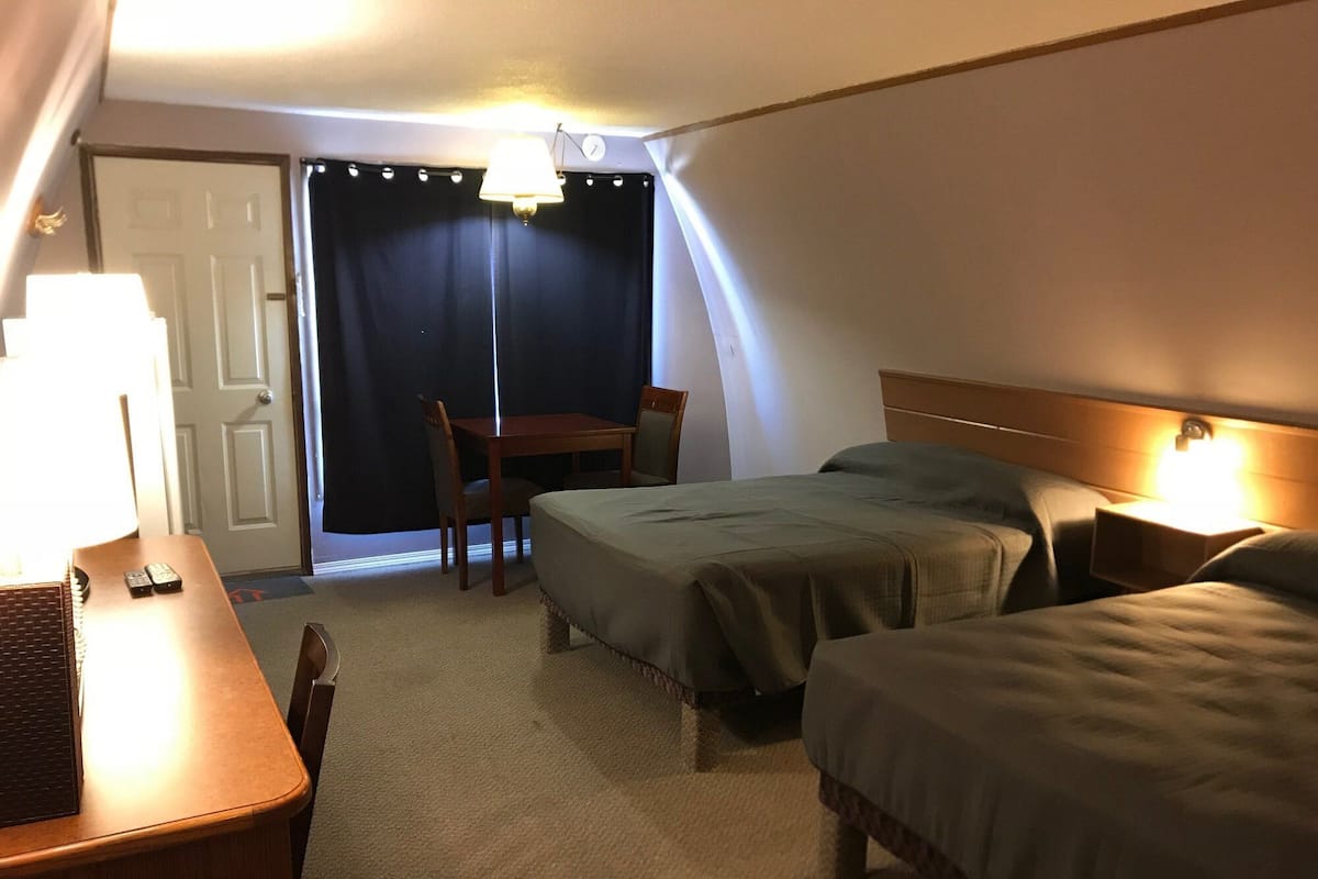 A hotel room with two beds, a desk with a lamp, a chair, and a table.