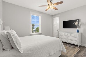 Ein Schlafzimmer mit Bett, Deckenventilator, Fenster mit Jalousien, Kommode und Flachbildfernseher.