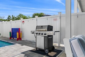 Ein tragbarer Grill mit mehreren Brennern und einem Seitentisch, in der Nähe eines Schwimmbeckens mit weißem Zaun und einem Spielbereich für Kinder im Hintergrund.