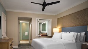 A hotel room with a bed, a desk, a chair, a ceiling fan, a bathroom with a mirror, and a patterned wall.