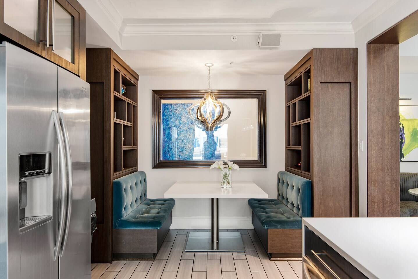 Une cuisine moderne dotée d’un réfrigérateur en acier inoxydable encastr é, d’une table à manger blanche avec deux bancs bleus à assise rembourrée et de meubles de rangement en bois.