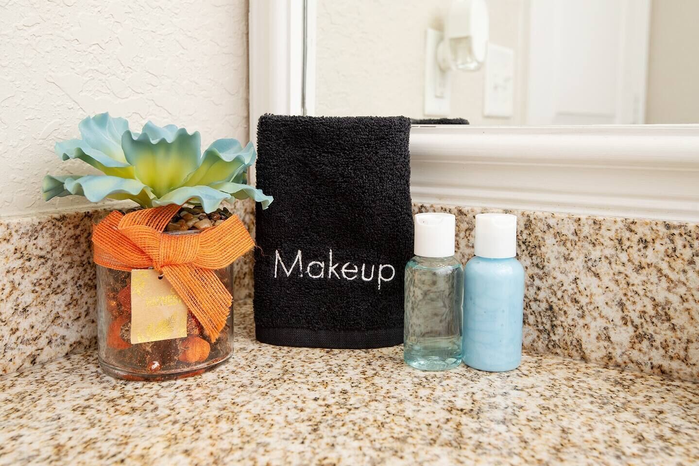 A bathroom counter with a black towel labeled "Makeup," a clear glass jar with an orange ribbon, and two bottles of liquid.
